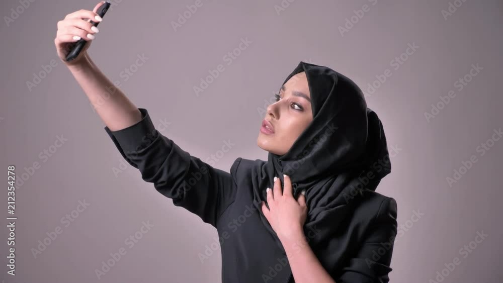 Young beautiful muslim girl in hijab is making selfie on her smartphone, communication concept, grey background Young beautiful muslim girl in hijab is making selfie on her smartphone, communication concept, grey background