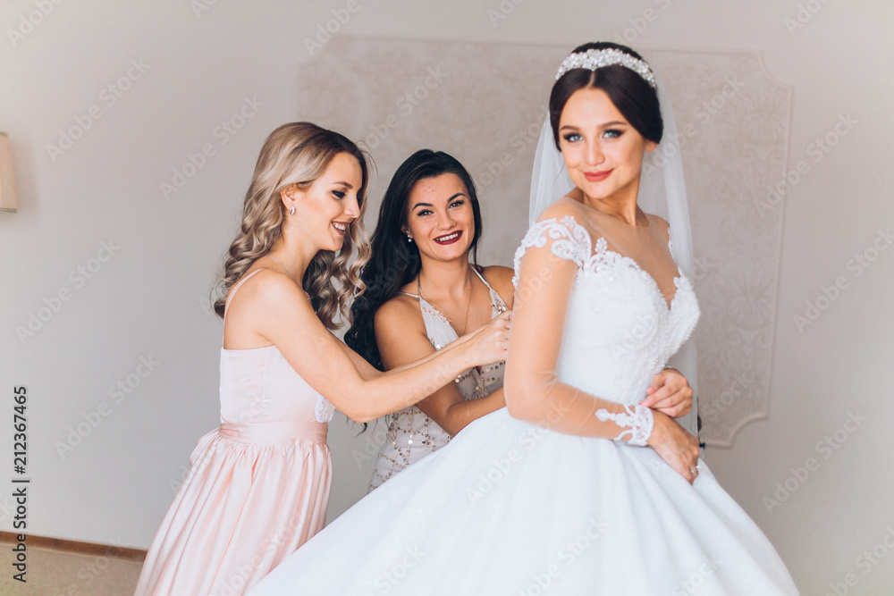 Bride preparation before wedding ceremony. Pleasant young bridesmaids helping the bride to get ready