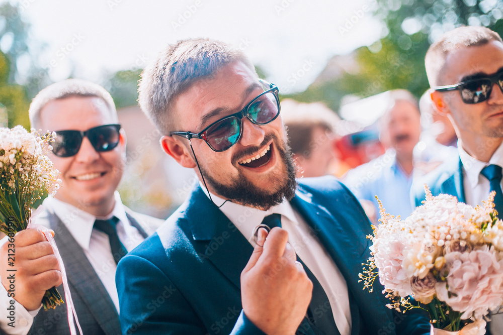 Groom and groomsmen have fun and walking to the restaurant. Wedding moment after ceremony. Guys in sunglasses and suite. Smiling wedding guests.