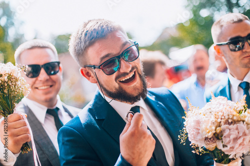 Groom and groomsmen have fun and walking to the restaurant. Wedding moment after ceremony. Guys in sunglasses and suite. Smiling wedding guests. photo