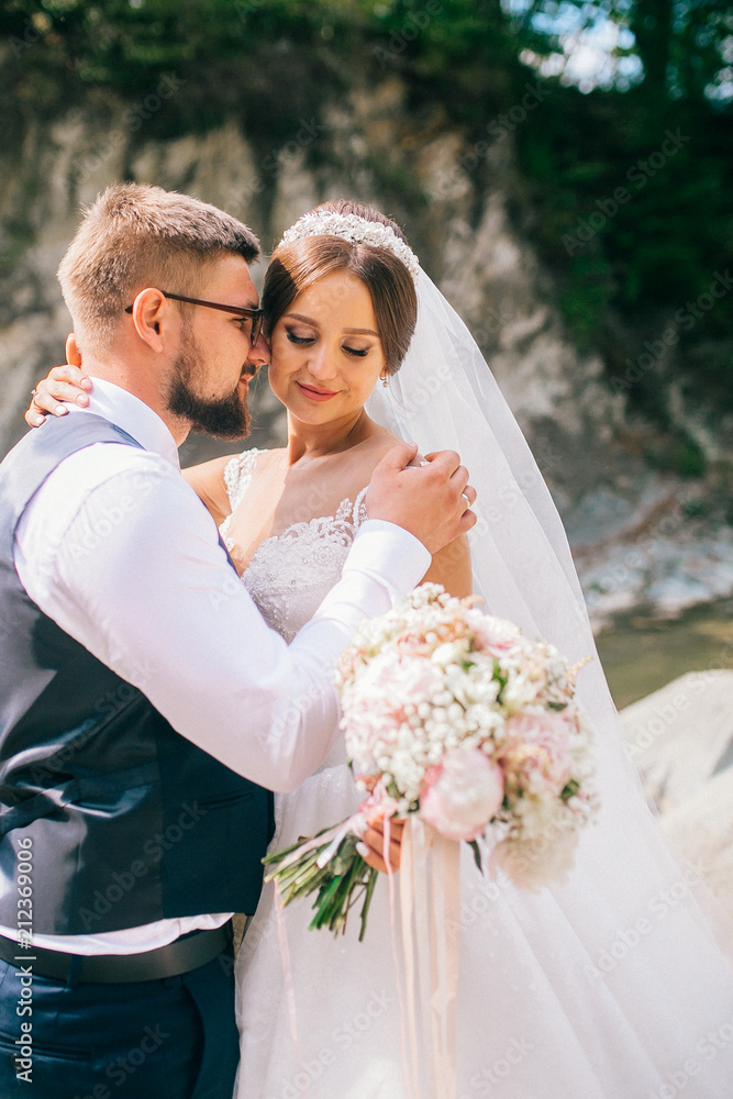 Couple newlyweds bride and groom kiss laughs and smiles to each other, happy and joyful moment. Man and woman in wedding clothes sit on the rock background mountain river. Wedding tender moments. Couple newlyweds bride and groom kiss laughs and smiles to each other, happy and joyful moment. Man and woman in wedding clothes sit on the rock background mountain river. Wedding tender moments.