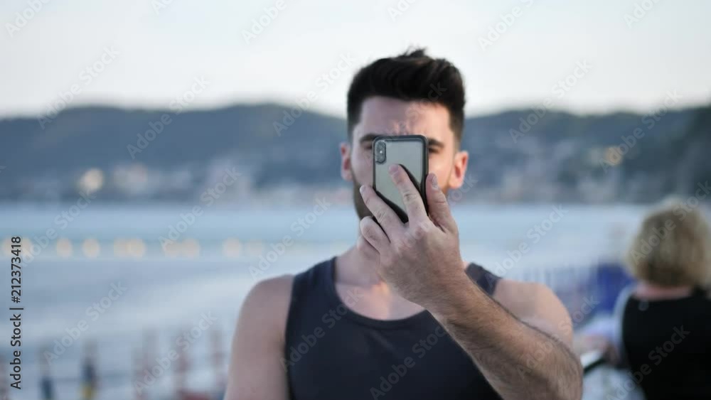 Young shirtless muscular handsome man doing videochat with smartphone by the sea at sunset Young shirtless muscular handsome man doing videochat with smartphone by the sea at sunset