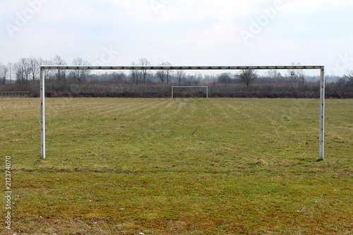 Metal soccer football goal posts without net on large football field with cut grass left to dry and small dried forest vegetation in background on cold winter day