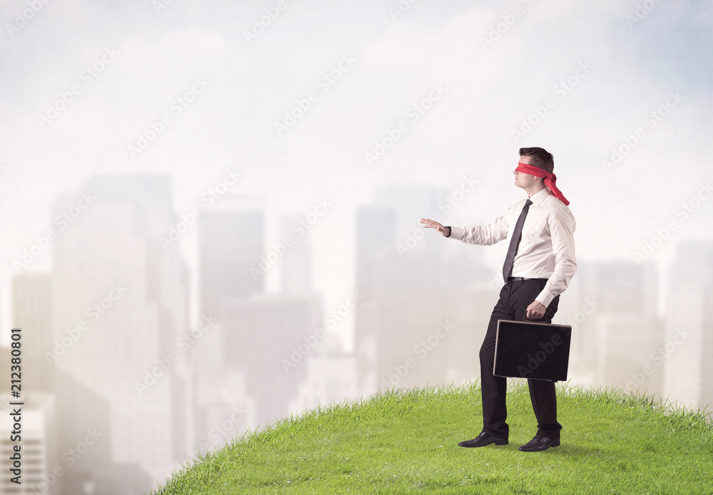 Young blindfolded businessman steps on a a patch of grass with a city in the background Young blindfolded businessman steps on a a patch of grass with a city in the background