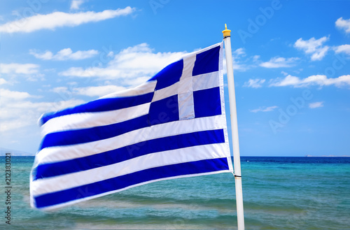 Greek flag waving in wind in blue sky and white clouds and sea waves around