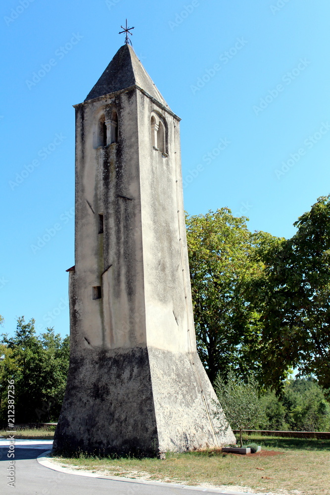 Leaning old stone church bell tower surrounded with trees and road under clear blue summer sky