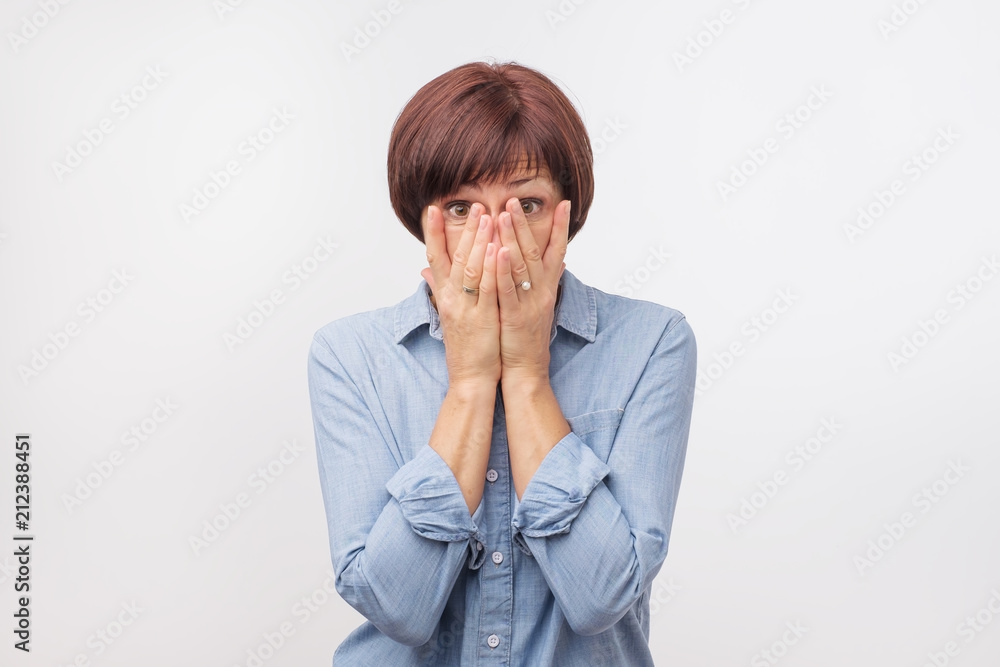 Close up portrait of attractive, mature woman in blue shirt closing her face with fingers. She is worried. She can't say anything. Getting bad news or hear gossip Close up portrait of attractive, mature woman in blue shirt closing her face with fingers. She is worried. She can't say anything. Getting bad news or hear gossip