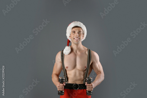 Young muscular man in Santa hat with dumbbells on gray background