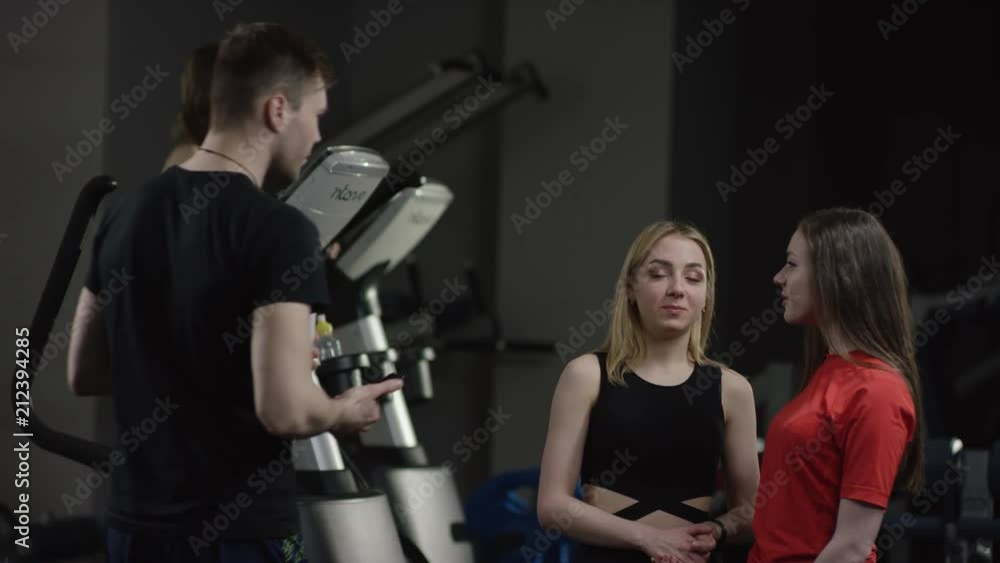 coach comes to greeting to beautiful girls in new gym and ask about them with girl engaged in simulators in the background coach comes to greeting to beautiful girls in new gym and ask about them with girl engaged in simulators in the background