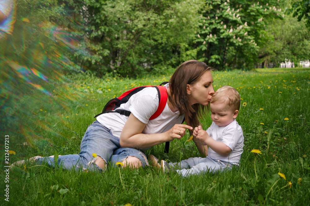 Family. Son and mother outdoors. A pregnant girl with a backpack is sitting on the lawn and kissing a little boy. He sits on the lawn and plays with a yellow dandelion. Blur effect Family. Son and mother outdoors. A pregnant girl with a backpack is sitting on the lawn and kissing a little boy. He sits on the lawn and plays with a yellow dandelion. Blur effect