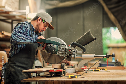 Young brunette man in work clothes by profession carpenter builder saws with a circular saw a wooden board on a wooden table in the workshop