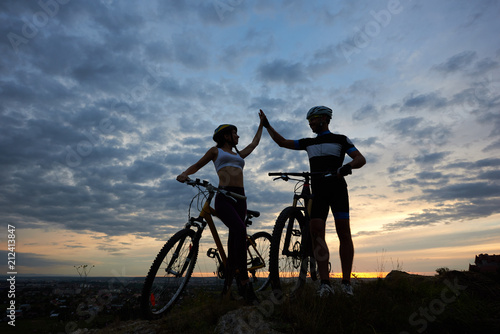 Silhouettes of two cyclists highing five after riding bikes. Sporty man and woman standing near bicycles and posing in twilight. Concept of motivation, sport and activities outdoors.