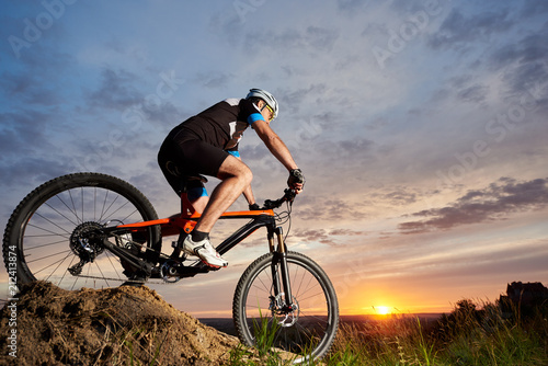 Active male cyclist wearing sportswear and helmet, riding bicycle alone and rolling down hill. Sporty and robust man cycling against beautiful sunset and rose-blue sky in the evening background