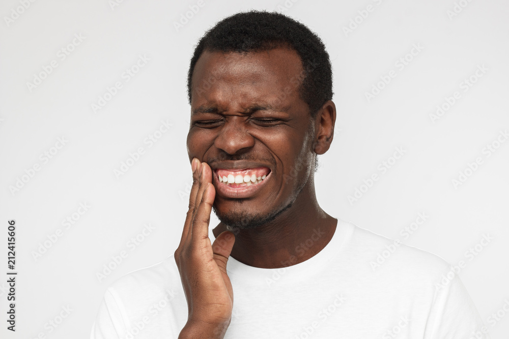 Indoor portrait of young worried african american man isolated on gray background, dressed in white t shirt, experiencing strong toothache pressing hand to chin because of sore jaws Indoor portrait of young worried african american man isolated on gray background, dressed in white t shirt, experiencing strong toothache pressing hand to chin because of sore jaws