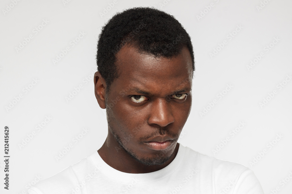 Close up portrait of young african american man in blank white t shirt isolated on gray background, looking at camera with anger