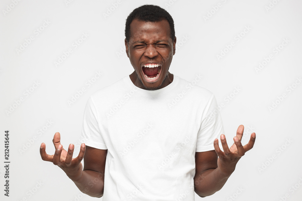 Handsome african american man in blank white t shirt, standing isolated against grey backgroung and screaming desperately, gesturing with both hands Handsome african american man in blank white t shirt, standing isolated against grey backgroung and screaming desperately, gesturing with both hands