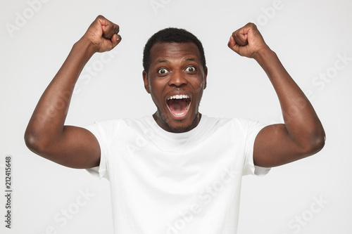 Young african american man in blank white t shirt, shouting while his team win, raised both fist in victory gesture, isolated on gray background