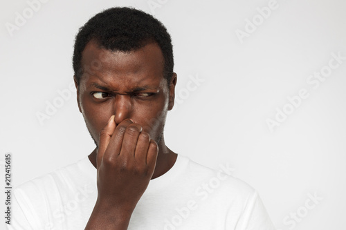 African american man in blank white tshirt, isolated on gray background holding his nose trying to escape from stinky smell and looking aside with worry looking for source of odor