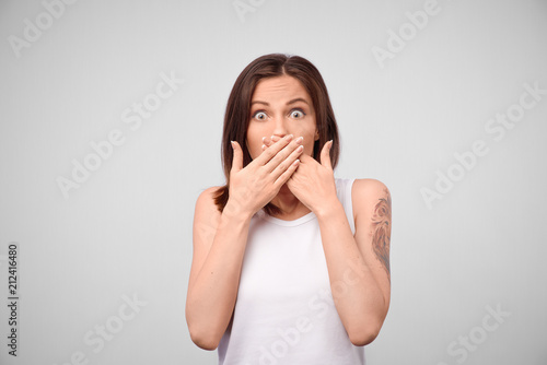 Portrait of a surprised young woman covering mouth with hands isolated over white background. Young woman covers mouth with hand, not tell, isolated on a gray background