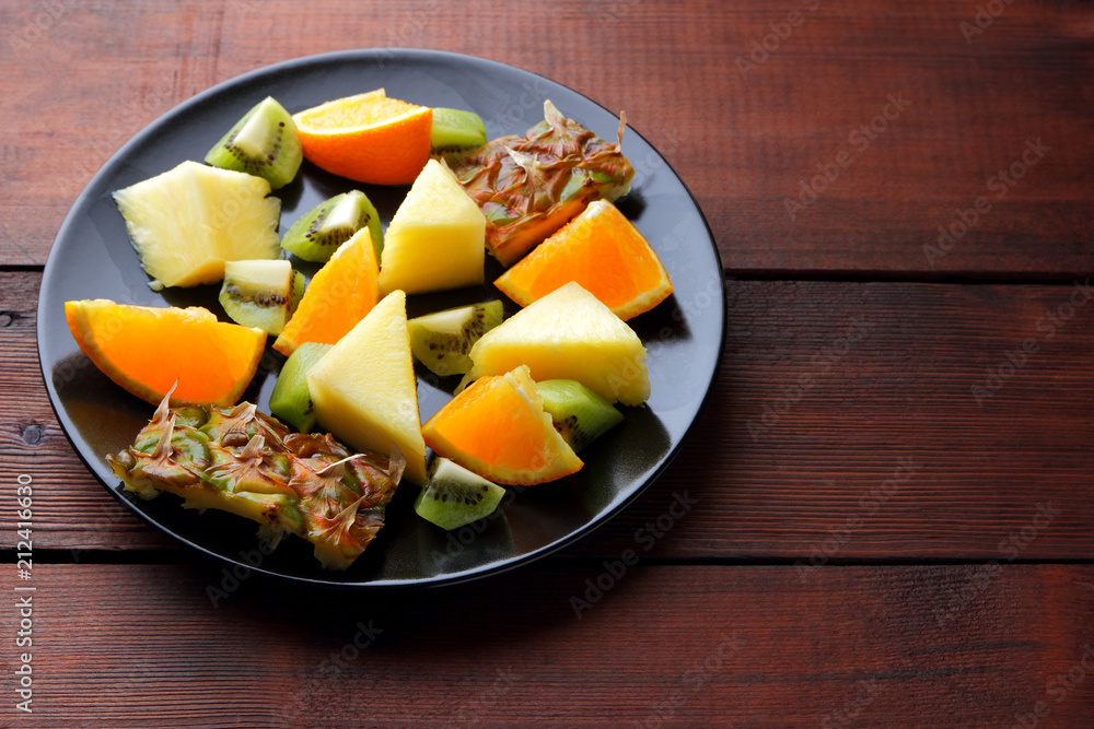 Fruit salad for vegans, pieces of pineapple, oranges and kiwi on a black plate, tropical fruits on wooden boards, citrus salad top view, vegetarian food Fruit salad for vegans, pieces of pineapple, oranges and kiwi on a black plate, tropical fruits on wooden boards, citrus salad top view, vegetarian food