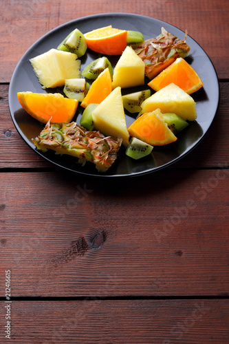 Fruit salad for vegans, pieces of pineapple, oranges and kiwi on a black plate, tropical fruits on wooden boards, citrus salad top view, vegetarian food