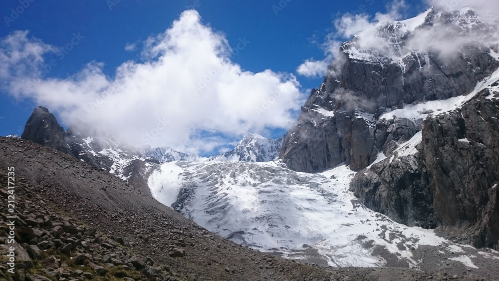 A glacier among the rocks, above it a blue sky and a white cloud.