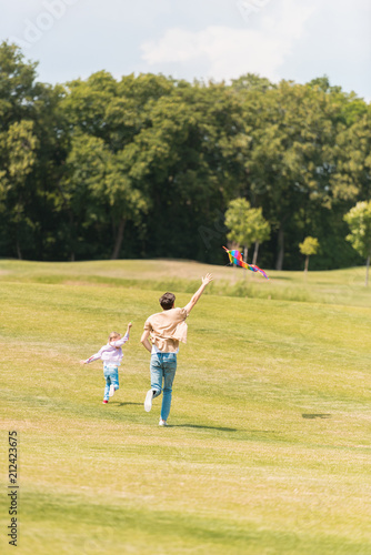 back view of father and daughter running on meadow and playing with kite