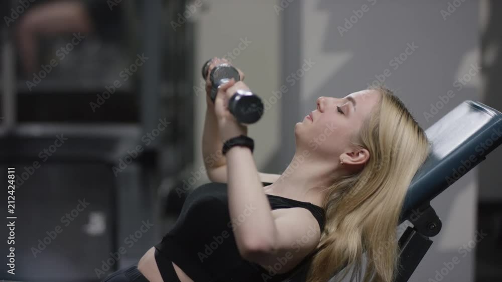 pretty blonde girl with straight hair in a black sport wear with blue eyes does a dumbbell press on an incline bench and coach comes up to fix her pretty blonde girl with straight hair in a black sport wear with blue eyes does a dumbbell press on an incline bench and coach comes up to fix her
