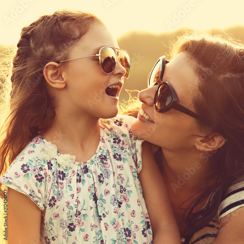 Happy fashion kid girl speaking to her mother in trendy sunglasses on nature sunset background. Closeup portrait of happiness.