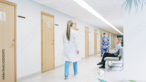 Fotografia In the Hospital Hallway, Busy Professional Personnel Walking by, Nurse Pushes Senior Man in the Wheelchair, Patients wait for their Doctor