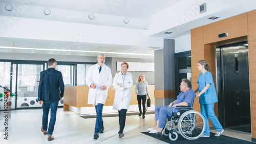 First Floor of the Busy Hospital, Doctors, Nurses and Personnel Busy Working, Assistant Moves Elderly Man in the Wheelchair. New Modern Medical Hospital with Professional Staff.