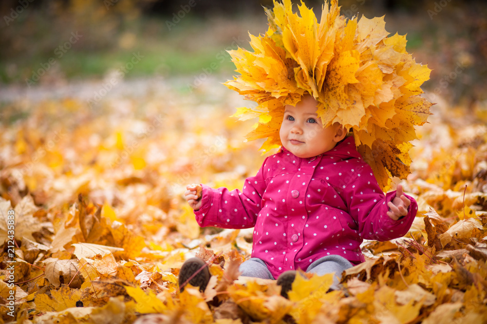 Little kid is playing and sitting in fallen leaves in autumn park. Baby is in big wreath of leaves. Girl is dressed in warm hat, jacket. - obrazy, fototapety, plakaty 
