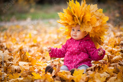 Little kid is playing and sitting in fallen leaves in autumn park. Baby is in big wreath of leaves. Girl is dressed in warm hat, jacket. photo