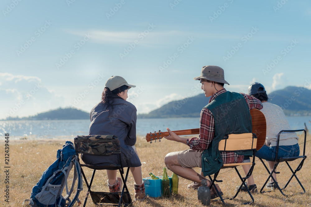 Camping group of friends asian people together, teenagers travel with backpack,tent man playing guitar song relaxing on vacation time holiday sitting on chairs near river and mountain view. Camping group of friends asian people together, teenagers travel with backpack,tent man playing guitar song relaxing on vacation time holiday sitting on chairs near river and mountain view.