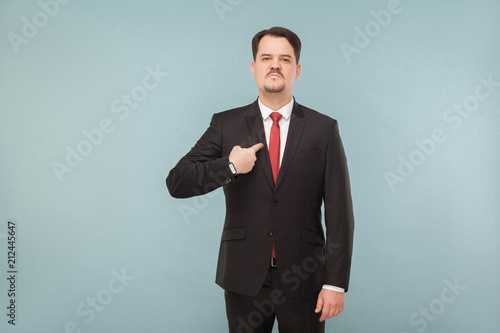 Man in classic wear pointing himself at camera. I did it and I'm proud of it. indoor studio shot. isolated on light blue background. handsome businessman with black suit, red tie and mustache.