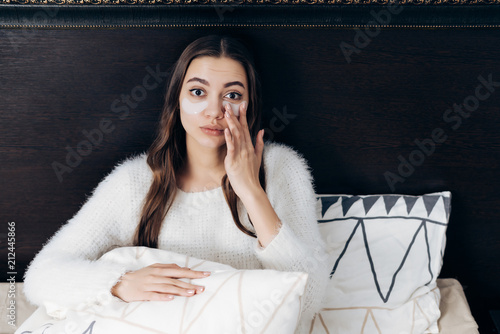 a wiry sleepy girl sits in a bed under a white blanket, under the eyes of a pachi against swelling photo