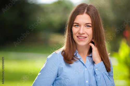 Close-up portrait - beautiful young brown-haired woman in a blue shirt looks at the camera on a background of blurred green trees and a lawn in the park.
