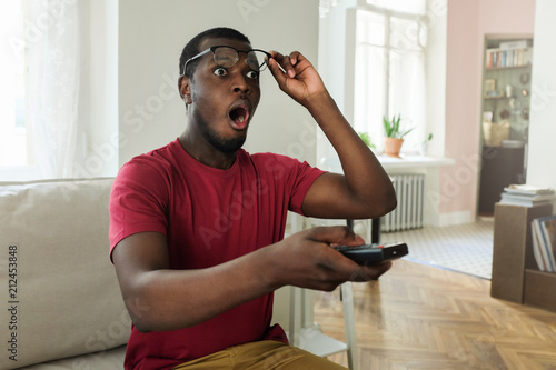 Horizontal picture of young African man spending free time at home watching television, looking surprised and shocked with open mouth raising eyeglasses to make sure what he sees on screen is real