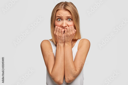 Nervous beautiful female bites fingernails, feel anxious, afraids of facing destiny, has scared expression, isolated over white background, worries someone will know her secret. Concerned woman