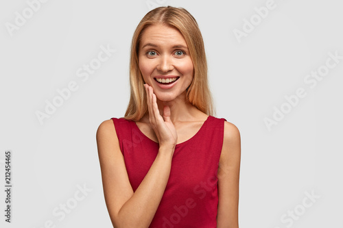 Joyful young pretty European female with light straight hair, being in good mood, looks happily at best friend who presents expensive present, wears casual red vest, isolated over white studio wall