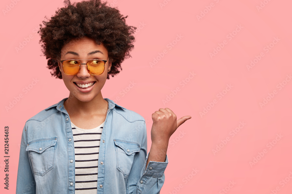 Studio shot of happy dark skinned African American female has crisp hair, toothy smile, points aside with thumb, stands indoor against pink background. People, advertisement, ethnicity concept Studio shot of happy dark skinned African American female has crisp hair, toothy smile, points aside with thumb, stands indoor against pink background. People, advertisement, ethnicity concept