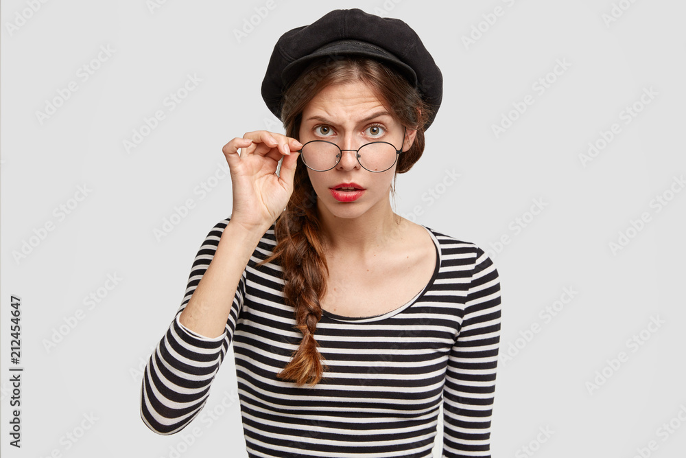 Attentive strict female teacher of French looks scrupulously through eyewear, wears striped sweater and beret, poses against white background. People, emotions and facial expressions concept Attentive strict female teacher of French looks scrupulously through eyewear, wears striped sweater and beret, poses against white background. People, emotions and facial expressions concept