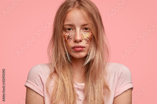 Headshot of beautiful woman with glitter on face and hair, has professional make up, looks seriously, contemplates about something, takes care of appearance, stands over pink background. Monochrome