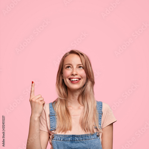 Happy young cute female teenager in denim overalls, indicates up, has positive smile, feels happy while notices something great upwards, isolated over pink background. Youth and advertisement