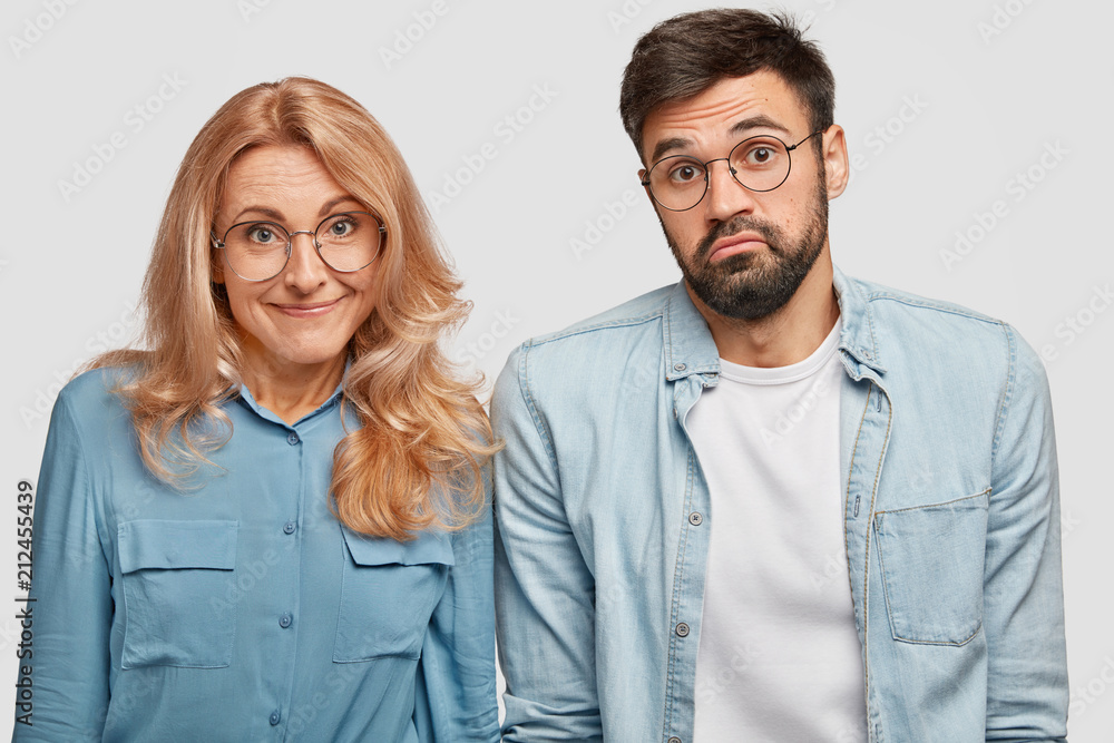 Horizontal shot of hesitant doubtful female and male colleagues, don`t know when to start work, stand next to each other, have clueless expressions, wear spectacles, isolated over white background Horizontal shot of hesitant doubtful female and male colleagues, don`t know when to start work, stand next to each other, have clueless expressions, wear spectacles, isolated over white background