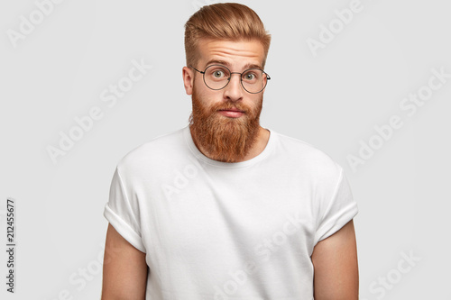 Puzzled ginger male freelancer feels surprised, looks in bewilderment, wears round spectacles, can`t undersatnd his duties, isolated over white background. People, reaction and style concept © wayhome.studio 