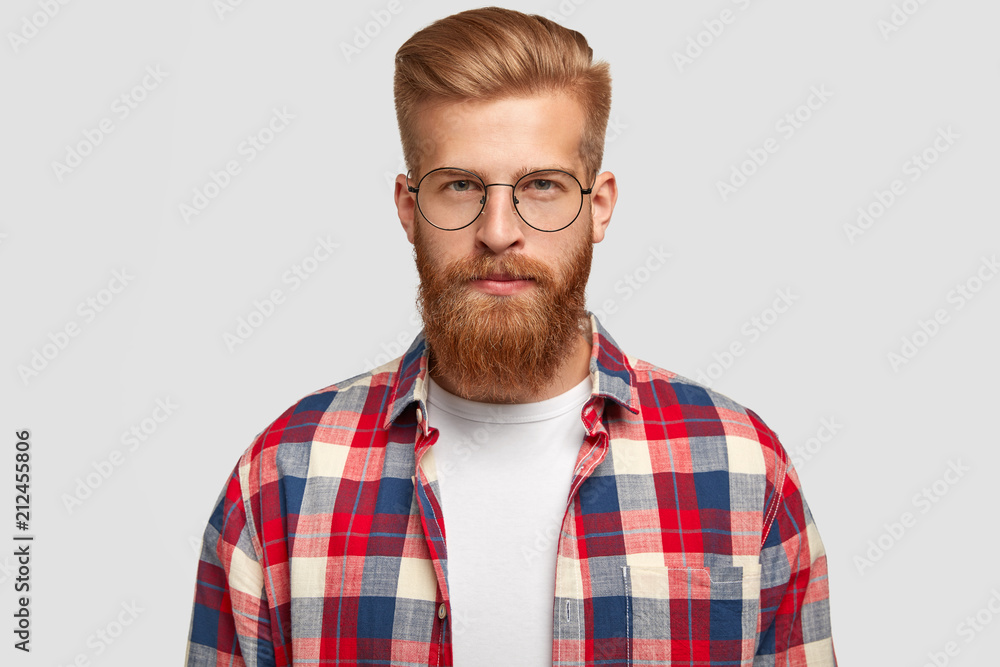 Fototapeta premium Studio shot of ginger male hipster in eyewear and checkered shirt, looks serious with confident facial expression, recieves necessary information, isolated over white background. People and style
