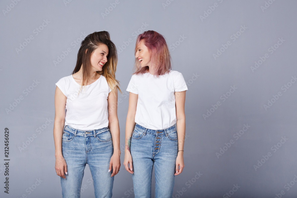 Horizontal portrait of two pretty women with happy expression, wears similar casual white t-shirt with denim, poses over gray background. Adorable lovely female student smile at each other. Copyspace Horizontal portrait of two pretty women with happy expression, wears similar casual white t-shirt with denim, poses over gray background. Adorable lovely female student smile at each other. Copyspace