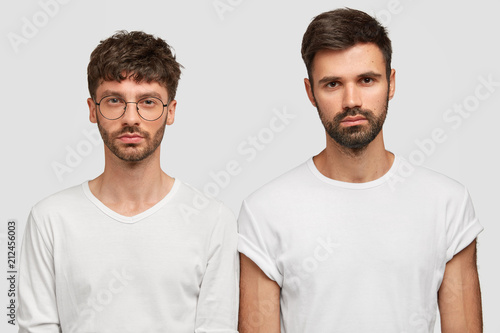 Indoor shot of serious two Caucasian men look directly at camera, wear casual clothes, have stubbles, work as friendly team, stand against white background. People, facial expressions concept