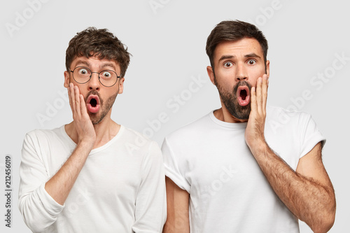 Surprised bearded male friends recieve shocking news, keep hands on cheeks, stare with eyes popped out, wears white clothes in one tone with background. People, emotions, astonishment concept
